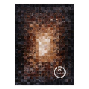 May include: A brown and black patchwork cowhide rug with a lighter brown center. The rug is made of squares of leather sewn together. The rug is made by Prime Cowhides.