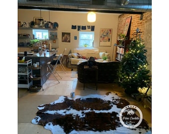 May include: A living room with a brown leather sofa, a white sofa, a small Christmas tree with white lights, and a cowhide rug. The room has exposed brick walls and a large window.