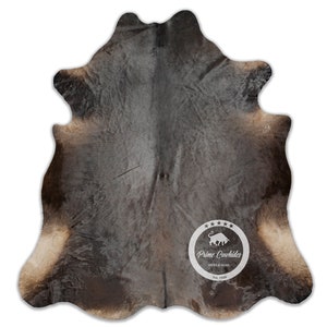 以下が含まれることがあります： 中央に「Prime Cowhides」のロゴが入った茶色と白の牛革の敷物。敷物は自然で素朴な外観と感触です。