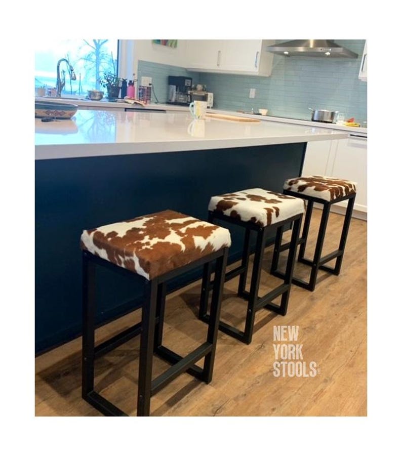Puede incluir: Tres taburetes de bar con marcos de metal negro y asientos rectangulares tapizados en piel de vaca marr&oacute;n y blanca. El texto NEW YORK STOOLS es visible en la esquina inferior derecha. Cada taburete cuenta con una base robusta.