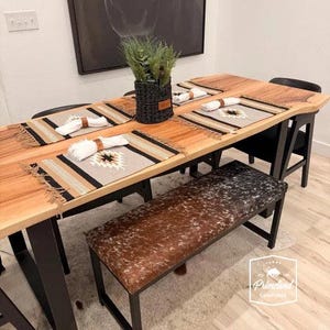 Puede incluir: Un conjunto de comedor con mesa de madera de borde natural y patas de metal negro, un banco tapizado en piel de vaca marrón y blanca, y sillas negras. La mesa está puesta con manteles individuales tejidos, servilletas blancas y una cesta.