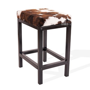 May include: A rectangular stool with a black metal frame and a seat upholstered in brown and white cowhide. The frame features visible bolt heads at the corners. The text "URK STOOLS" is printed in white on the corner of the cowhide seat.