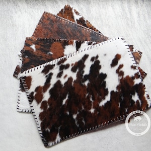 May include: A stack of four cowhide coasters with a brown and white pattern. The coasters are stitched with a black thread and have a brown leather border. The coasters are made by Prime Cowhides, Est. 1988.