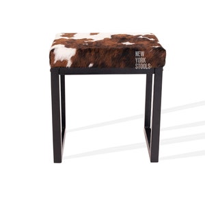 May include: A black metal frame stool with a brown and white cowhide seat. The stool has a square top and four legs. The seat has the text "NEW YORK STOOLS" printed on it.