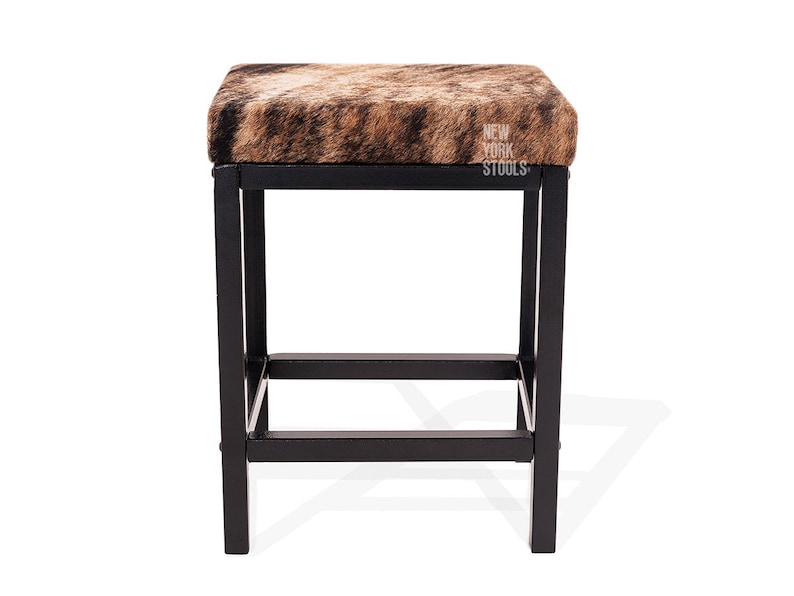 Puede incluir: Un taburete cuadrado con asiento de piel con estampado animal marr&oacute;n y negro y estructura de metal negro. El asiento tiene el texto blanco NEW YORK STOOLS impreso en el lado derecho. La base incluye cuatro patas rectas unidas por barras.