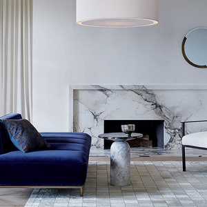 K&ouml;nnte beinhalten: Ein Wohnzimmer-Interieur mit einem blauen Samtsofa, einem wei&szlig;en Sessel, einem Marmorkamin und einem runden, wei&szlig;en Beistelltisch. Der Raum hat einen wei&szlig;en Teppich mit einem schwarz-wei&szlig;en Muster.