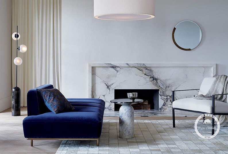 K&ouml;nnte beinhalten: Ein Wohnzimmer-Interieur mit einem blauen Samtsofa, einem wei&szlig;en Sessel, einem Marmorkamin und einem runden, wei&szlig;en Beistelltisch. Der Raum hat einen wei&szlig;en Teppich mit einem schwarz-wei&szlig;en Muster.