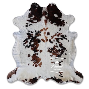 以下が含まれることがあります： 茶色と白の斑点模様が特徴的なカウハイドラグ。ラグは不規則な形状で、柔らかい質感です。ラグの中央にはPrimeland Cowhidesのロゴが見えます。
