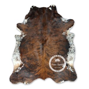 Peut inclure: Tapis en peau de vache brun et blanc avec un logo de taureau noir et blanc au centre. Le logo indique "Prime Cowhide Hides & Skins Est. 1999".