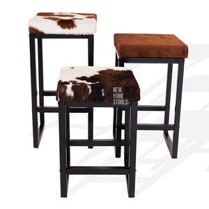 May include: Three black metal bar stools with brown and white cowhide seats. The stools are stacked on top of each other. The middle stool has a label that reads "NEW YORK STOOLS".