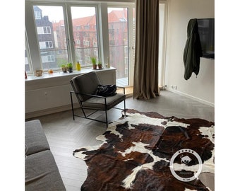 May include: A room interior with a cowhide rug in brown and white. A grey armchair with a black frame is positioned near a large window. The room has wooden flooring and neutral walls. A black bag is on the chair. The room has a modern design.
