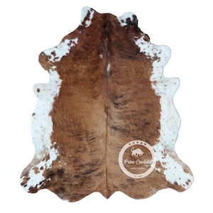 Puede incluir: Una alfombra de piel de vaca con un patrón natural de manchas marrones y blancas. La alfombra tiene una forma irregular y un aspecto suave y texturizado. Un logotipo de marca es visible en el centro.