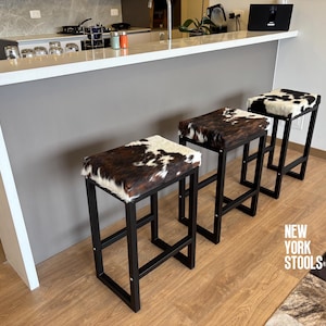 Puede incluir: Tres taburetes de bar con marcos de metal negro y asientos rectangulares tapizados en piel de vaca marrón, negra y blanca. Los taburetes presentan un diseño geométrico limpio. El texto NEW YORK STOOLS es visible en la esquina inferior derecha.