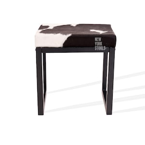 May include: Black and white cowhide upholstered stool with a black metal frame. The stool has the text "NEW YORK STOOLS" printed on the top.