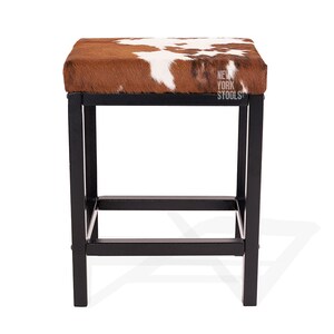 Puede incluir: Un taburete cuadrado con un coj&iacute;n de asiento de piel de vaca marr&oacute;n y blanca sobre una estructura de metal negro con cuatro patas y una barra de soporte inferior. El texto NEW YORK STOOLS est&aacute; impreso en blanco en el lado derecho del asiento.