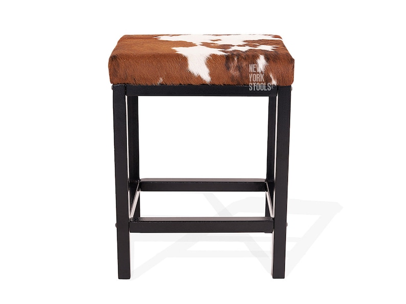 Puede incluir: Un taburete cuadrado con un coj&iacute;n de asiento de piel de vaca marr&oacute;n y blanca sobre una estructura de metal negro con cuatro patas y una barra de soporte inferior. El texto NEW YORK STOOLS est&aacute; impreso en blanco en el lado derecho del asiento.