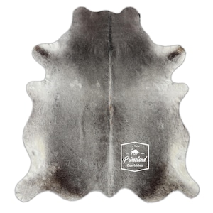 以下が含まれることがあります： 中央に「Primeland Cowhides」と書かれた白いロゴが付いた灰色の牛革の敷物です。