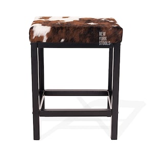 Puede incluir: Un taburete cuadrado con asiento de piel de vaca marr&oacute;n y blanca y estructura de metal negro. El asiento presenta el texto NEW YORK STOOLS impreso en blanco. La base consta de cuatro patas rectas conectadas por barras de soporte horizontales.