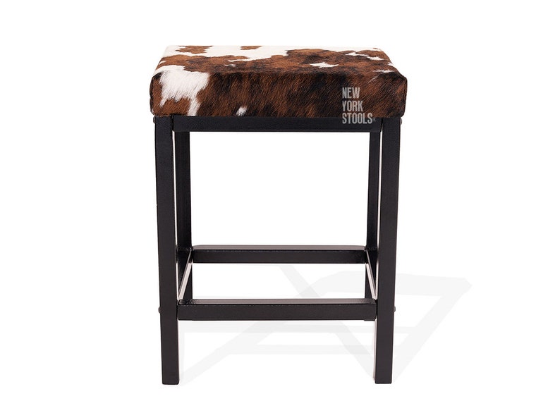 Puede incluir: Un taburete cuadrado con asiento de piel de vaca marr&oacute;n y blanca y estructura de metal negro. El asiento presenta el texto NEW YORK STOOLS impreso en blanco. La base consta de cuatro patas rectas conectadas por barras de soporte horizontales.