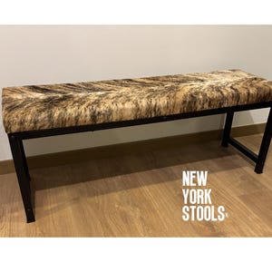 Puede incluir: Un banco largo y rectangular con un marco de metal negro y un asiento tapizado en piel de vaca marrón y blanco. El banco está sentado en un suelo de madera. El texto "NEW YORK STOOLS" es visible en el suelo.