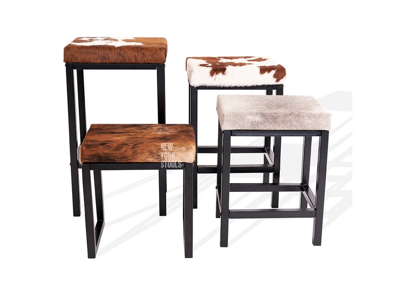 Puede incluir: Cuatro taburetes cuadrados con marcos de metal negro y asientos de piel de vaca en varios tonos de marr&oacute;n, blanco y gris. Un asiento presenta el texto NEW YORK STOOLS. Los taburetes est&aacute;n dispuestos a diferentes alturas con patrones.