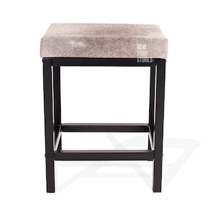 Puede incluir: Un taburete cuadrado con un asiento de piel animal texturizada en gris claro y blanco y una estructura de metal negro. El asiento presenta el texto blanco NEW YORK STOOLS impreso en el lado derecho. La base consta de cuatro patas rectas.
