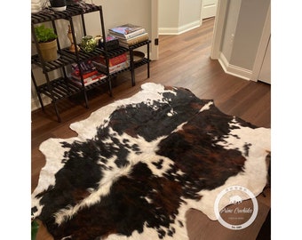 May include: A brown, black and white cowhide rug with a white background and brown and black patches. The rug is on a hardwood floor in a room with a wooden shelf unit in the background.