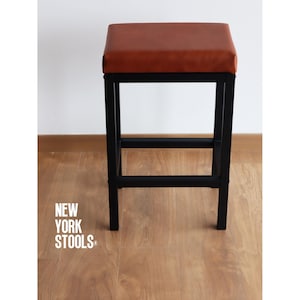 Puede incluir: Taburete cuadrado con asiento de cuero marrón y estructura de metal negro. El taburete tiene cuatro patas y una barra de soporte horizontal. El texto "NEW YORK STOOLS" es visible en el lado izquierdo de la imagen.