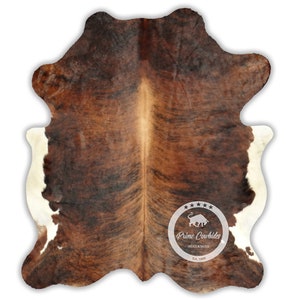 以下が含まれることがあります： 茶色と白の牛革の敷物で、自然な斑点模様があります。敷物は白い縁取りがあり、中央には「Prime Cowhides Hides & Rugs Est. 1999」と書かれたロゴがあります。