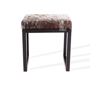 May include: A black metal stool with a brown and white cowhide seat. The stool has a simple, modern design and is perfect for adding a touch of rustic charm to any room. The stool is labelled "New York Stools".