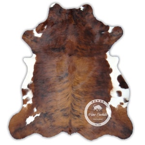 以下が含まれることがあります： 黒と茶色の模様が入った茶色と白の牛革の敷物。敷物には「Prime Cowhides」のロゴと、星5つの評価と「HIDES & SKINS Est. 1999」の文字が書かれています。