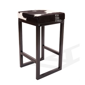 Op de afbeelding: Zwarte metalen barstoel met een vierkante zitting van koeienhuid. De zitting heeft een zwart-wit patroon. De stoel heeft een metalen frame en een zwarte afwerking. De stoel is gelabeld met "New York Stools".