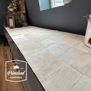 Può includere: Runner da tavolo rettangolare in pelle di mucca bianco sporco con un design patchwork. Il runner è composto da singoli quadrati di pelle di mucca cuciti insieme. Il logo Primeland Cowhides è visibile in basso a sinistra.