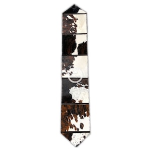 May include: A rectangular table runner with a cowhide pattern. The runner is made of rectangular panels of brown, black, and white cowhide, stitched together. The bottom edge is pointed. A circular tag is visible near the center.
