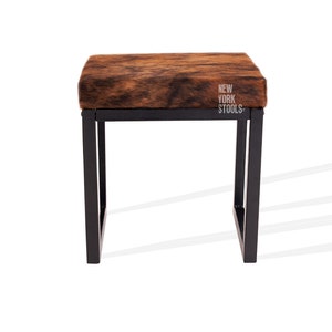 May include: A brown and black cowhide upholstered stool with a black metal frame. The stool has a square top and is approximately 46 cm tall. The stool is labelled "New York Stools".