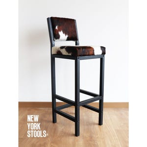 May include: Black metal bar stool with a cowhide seat. The stool has a simple, industrial design and is perfect for a modern kitchen or bar.