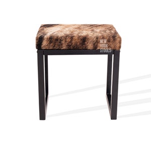 May include: A black metal stool with a brown and white cowhide seat. The stool has a simple, modern design and is perfect for adding a touch of rustic charm to any room. The stool is labelled "New York Stools".