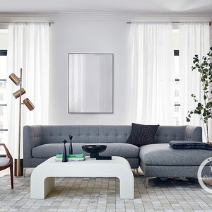 K&ouml;nnte beinhalten: Ein graues Ecksofa mit einem wei&szlig;en Couchtisch und einem wei&szlig;en B&uuml;cherregal in einem modernen Wohnzimmer. Das Sofa hat eine graue Wolldecke und ein schwarz-wei&szlig; gemustertes Kissen. Der Couchtisch hat eine Schale und B&uuml;cher darauf. Das B&uuml;cherregal hat B&uuml;cher und eine Pflanze darin.