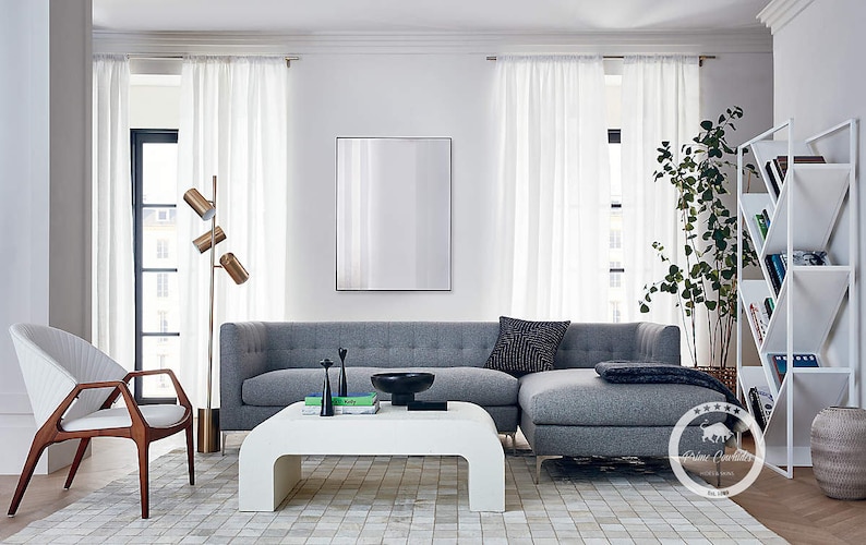 K&ouml;nnte beinhalten: Ein graues Ecksofa mit einem wei&szlig;en Couchtisch und einem wei&szlig;en B&uuml;cherregal in einem modernen Wohnzimmer. Das Sofa hat eine graue Wolldecke und ein schwarz-wei&szlig; gemustertes Kissen. Der Couchtisch hat eine Schale und B&uuml;cher darauf. Das B&uuml;cherregal hat B&uuml;cher und eine Pflanze darin.