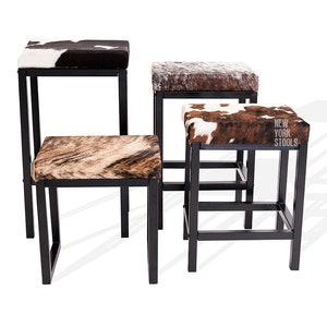 May include: Four black metal bar stools with different cowhide patterned seats. The stools are arranged in a staggered formation. The seat of the stool in the front has the text "NEW YORK STOOLS" printed on it.