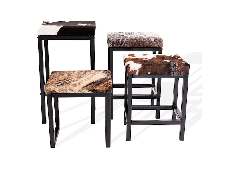 Puede incluir: Cuatro taburetes con marcos de metal negro y asientos cuadrados tapizados con varios patrones de piel de vaca. Los asientos presentan texturas de pelaje marr&oacute;n, blanco y negro. Un taburete muestra el texto NEW YORK STOOLS en el lateral.