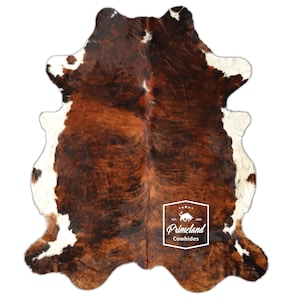 Puede incluir: Una alfombra de piel de vaca con un patrón natural de color marrón, blanco y negro. La alfombra tiene una forma irregular y una textura suave. El logotipo de Primeland Cowhides es visible en el centro inferior de la alfombra.