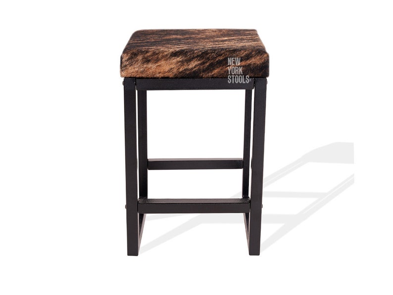 Puede incluir: Un taburete cuadrado con asiento de piel de vaca marr&oacute;n y negra y estructura de metal negro. El asiento presenta el texto blanco NEW YORK STOOLS impreso en el lado derecho. El taburete tiene cuatro patas rectas unidas por barras.