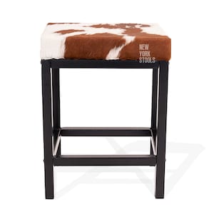 Puede incluir: Un taburete cuadrado con asiento de piel de vaca marr&oacute;n y blanca y estructura de metal negro. El asiento presenta el texto NEW YORK STOOLS impreso en blanco en el lado derecho. La base consta de cuatro patas rectas unidas por barras horizontales.