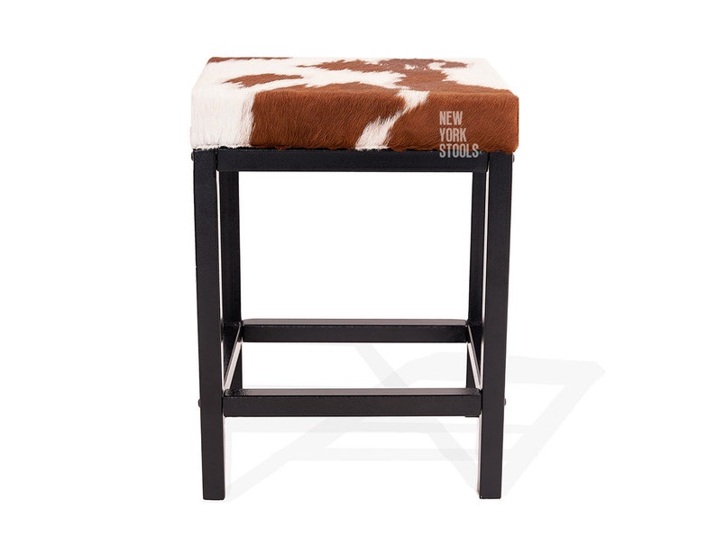 Puede incluir: Un taburete cuadrado con asiento de piel de vaca marr&oacute;n y blanca y estructura de metal negro. El asiento presenta el texto NEW YORK STOOLS impreso en blanco en el lado derecho. La base consta de cuatro patas rectas unidas por barras horizontales.