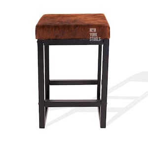 Puede incluir: Un taburete cuadrado con asiento de piel texturizada marr&oacute;n y estructura de metal negro. El asiento presenta el texto blanco NEW YORK STOOLS impreso en la esquina superior derecha. La estructura consta de cuatro patas rectas con travesa&ntilde;os.