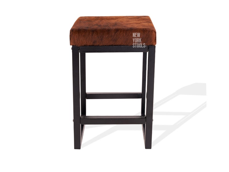 Puede incluir: Un taburete cuadrado con asiento de piel texturizada marr&oacute;n y estructura de metal negro. El asiento presenta el texto blanco NEW YORK STOOLS impreso en la esquina superior derecha. La estructura consta de cuatro patas rectas con travesa&ntilde;os.
