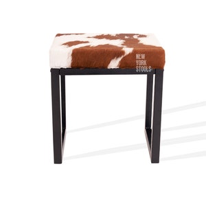 May include: A brown and white cowhide upholstered stool with a black metal frame. The stool has the text "NEW YORK STOOLS" printed on the top.