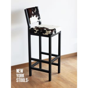 Puede incluir: Taburete de bar de metal negro con un asiento de piel de vaca blanca y negra. El taburete tiene un diseño simple e industrial, perfecto para una cocina o bar moderno.