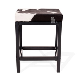 Puede incluir: Un taburete cuadrado con un asiento de patr&oacute;n de piel de vaca en blanco y negro y una estructura de metal negro. El asiento presenta el texto NEW YORK STOOLS impreso en blanco en el lado derecho. La estructura tiene cuatro patas rectas.